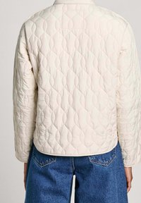 Prošívaná béžová bunda s texturovaným vzorem, kulatým límcem a širokými rukávy, doplněná modrými džínovými kalhotami.
