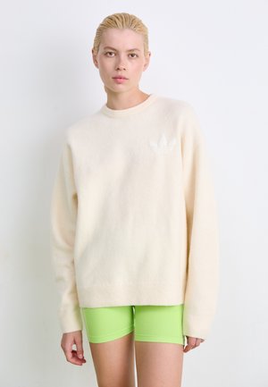 Pull crème avec un ourlet et des poignets côtelés ; arbore un logo Adidas blanc subtil sur la poitrine gauche, associé à un short vert vif.