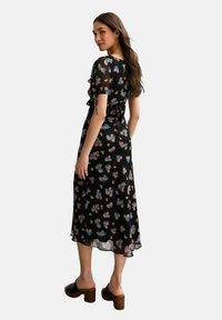 Robe noire à fleurs avec des manches courtes à volants, présentant une taille cintrée et un ourlet fluide. Accentuée par des fleurs bleues et roses. Portée avec des mules à talons.