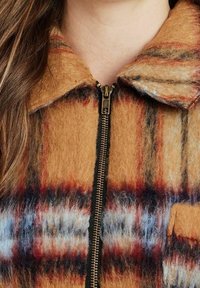 Veste à fermeture éclair à carreaux marron et orange, en tissu doux et texturé, avec un col et des accents bleus contrastants partout.