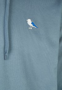 Felpa leggera blu con cappuccio, caratterizzata da un piccolo uccello ricamato in bianco e blu sul lato sinistro del petto. Il materiale sembra morbido e tessurizzato.