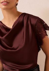 Top din satin bordeaux cu decolteu drapat și mâneci scurte din dantelă, având o textură fină și o drapare elegantă. Centura neagră este vizibilă.