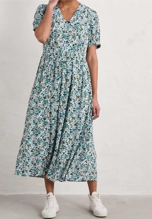 Frau trägt ein ärmelloses, knielanges Kleid mit blauen, grünen und weißen Blumenmustern, kurzen Puffärmeln und weißen Turnschuhen, steht vor einer schlichten Wand.