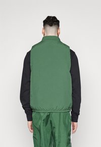 Man med flätat hår som bär en grön utanpåliggande jacka över en svart långärmad tröja och gröna glänsande byxor, vänd bort från kameran.