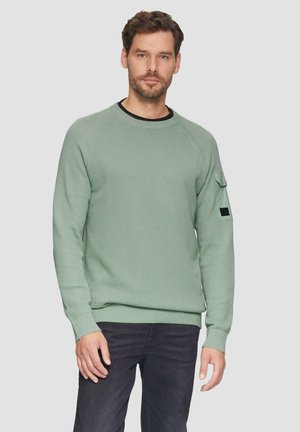 Pull en maille verte texturé avec des manches longues et un ourlet côtelé. Il présente des manches raglan et une petite poche sur le bras gauche. Porté par-dessus une chemise noire.