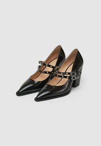 Scarpe con tacco in vernice nera, con punta affusolata, caratterizzate da doppie cinghie alla caviglia con borchie metalliche decorative e design con tacco a blocco.