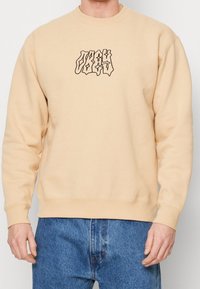 Světle béžová mikina s černým grafickým designem na přední straně, s kulatým výstřihem a žebrovanými manžetami a lemem. Nosit s modrými džíny.