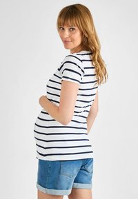 Materinski top ima mornarsko in belo horizontalno črtast vzorec, kratke rokave in prilegajočo obliko; je v paru s trajnimi denim kratkimi hlačami s zvitimi robovi.