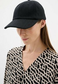 Black baseball cap with a textured fabric and stitched seams, worn by a person in a patterned black and white blouse.
