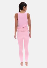 Mujer con el cabello trenzado, vistiendo una blusa sin mangas rosa claro con volantes y leggins a juego, de pie descalza y de espaldas.