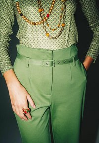 Green trousers with a belted waist and side pockets, paired with a patterned green top. Highlighted with layered colorful bead necklaces and a decorative ring.