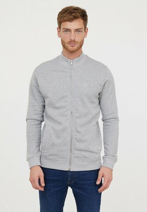 Sweatshirt gris zippé avec un col montant, des poignets et un ourlet côtelé, et un logo discret sur la poitrine. Porté avec un jean bleu foncé.