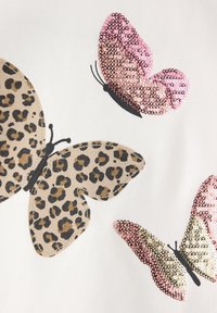 Tre design di farfalle su un tessuto bianco; uno con stampa leopardo e due decorati con paillettes rosa e oro.