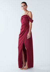 Večernja haljina boje burgundca s dizajnom bez ramena, naboranim detaljima, visokim prorezom i satenskom teksturom; uparena sa zlatnim sandalama s trakicama.