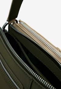 Grüne Lederhandtasche mit mehreren Reißverschlusstaschen. Silberne Beschläge, glatte Textur und kontrastierendes beiges Innenfutter sichtbar.