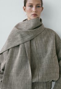 Donna che indossa una giacca grigia oversize con trama, con una larga sciarpa avvolta intorno al collo, su uno sfondo chiaro e uniforme.