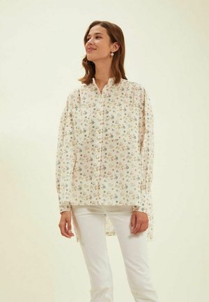Chemisier à manches longues et à boutons avec un motif floral, présentant un fond crème, des accents colorés et un ourlet arrière légèrement plus long.