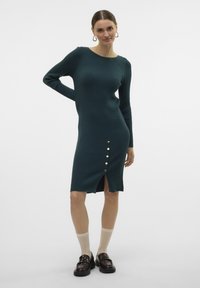 Ribota teal krāsas adīta kleita ar garām piedurknēm, priekšēju šķēlumu un sešām zelta pogām. Kombinēta ar melniem mugursomas apaviem un bežām sporta zeķēm.