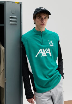 Tricou sport verde și negru cu mâneci lungi și guler cu fermoar, având logo-uri albe ale brandului și stema echipei. Purta de un model masculin lângă un dulap.