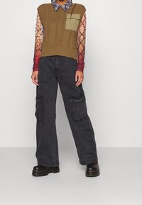 Brown knitted vest with cable pattern, over a multicolored snake print long-sleeve shirt, paired with gray cargo pants and black boots.