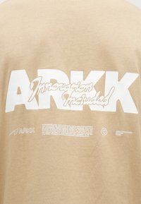 Bežová tkanina s velkým bílým textem "ARKK", italikou napsané "Inovace zahrnuta" jako překryv, a menším tištěným popisem a logy níže.