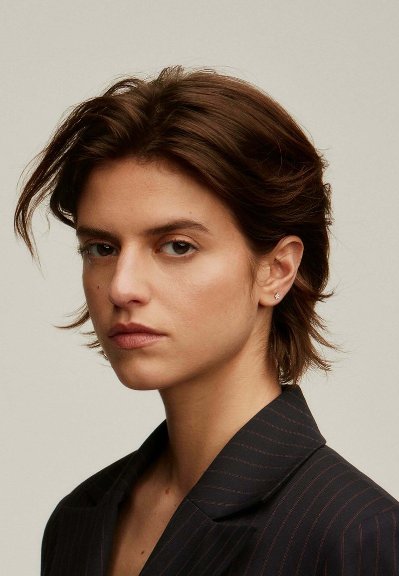 Retrato de una persona con cabello corto castaño, que lleva una chaqueta de traje oscura con finas rayas. Accesorios mínimos, textura facial sutil.