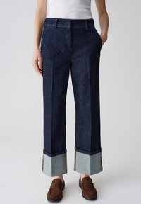 Pantalons en denim foncé à jambes larges avec un revers bleu clair, présentant un design classique à cinq poches et une coupe droite. Des mocassins marron complètent le look.