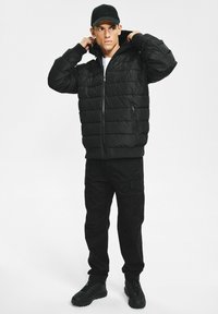 Czarna pikowana kurtka puchowa z pikowanym wzorem, zapięciem na zamek i ściągaczami przy mankietach. Na niej dopasowane czarne spodnie cargo i solidne czarne trampki.
