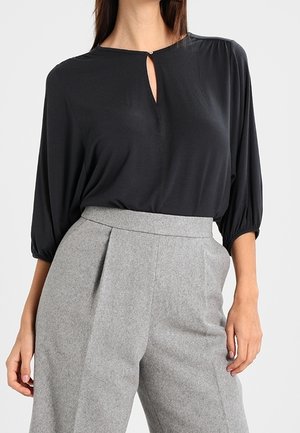 Blouse noire avec un décolleté en forme de trou de serrure, manches amples et léger brillant, assortie à un pantalon tailleur gris clair taille haute avec des plis.