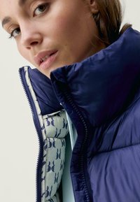 Mornarska puhovka z visokim ovratnikom, ki ima gladko zunanjost in vzorčeno svetlo modro podlogo. Zadrga z kontrastnim šivanjem.