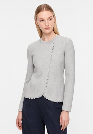 Donna che indossa una maglia a maniche lunghe grigio chiaro con texture e bordi smerlati, abbinata a pantaloni blu scuro, guarda verso sinistra.