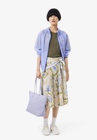 Chaqueta ligera azul claro y transparente sobre una camiseta oliva, combinada con una falda plisada de colores múltiples y estampada. Sosteniendo un bolso tote en lavanda suave. Zapatillas blancas.