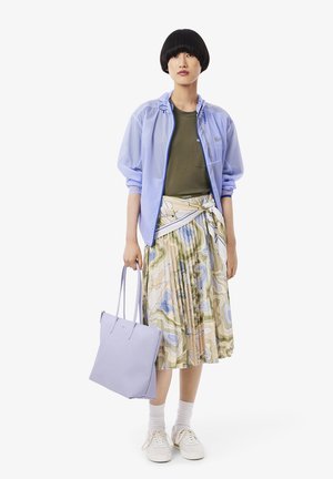 Lichtblauw, doorschijnend jasje over een olijfgroen T-shirt, gecombineerd met een geplooide, patroonrijke rok met meerdere kleuren. Houdt een zacht lavendelkleurige totebag vast. Witte sneakers.