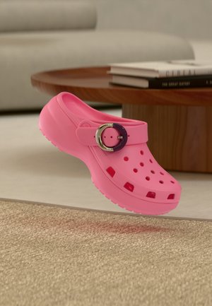 Leuchtend pinker Clog mit Belüftungslöchern und einer großen, runden Schnalle, schwebend über einem strukturierten beigen Teppich in der Nähe eines hölzernen Couchtisches mit Büchern.