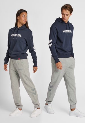 Mujer y hombre vistiendo sudaderas con capucha azul marino a juego y pantalones deportivos grises con zapatillas blancas, posando frente a un fondo claro y liso.