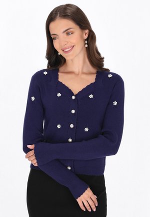 Vrouw die een marineblauwe cardigan draagt met parelknopen en bloemige parelversieringen, zacht glimlachend met gekruiste armen.