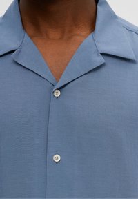 Kortärmad blå skjorta tillverkad av ett mjukt tyg; har en platt krage och en enda vit knapp vid halsen.