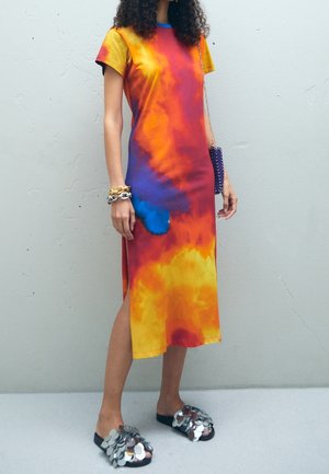 Dress features a multicolored tie-dye pattern with shades of orange, yellow, and blue. Short sleeves, side slit, and a relaxed fit. Black sandals with metallic accents.