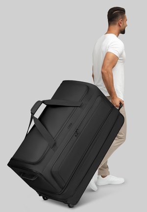 Sac de sport noir à roulettes avec une texture lisse, poignées latérales, poche zippée et une taille imposante, tiré par une personne en vêtements légers.