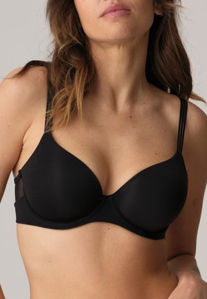 Mujer con un sujetador negro con aro y doble tira en los hombros, mostrada desde el cuello hasta el abdomen sobre un fondo liso.