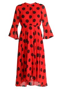Robe rouge à pois avec un design fluide, tissu transparent, manches longues évasées et taille froncée, ornée de grands motifs circulaires noirs.