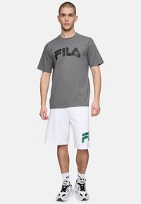 Camiseta gris de manga corta con un logo negro de FILA, combinada con pantalones cortos blancos que tienen un logo verde de FILA, y zapatillas deportivas en blanco y negro.