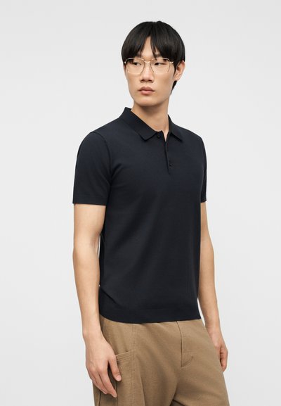 Jeune homme portant des lunettes à monture dorée, un polo noir à manches courtes et un pantalon beige, regardant vers sa gauche devant un fond uni.