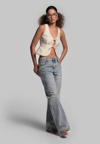 Cremfarbener, cropped Top mit Bindeband, kombiniert mit hellblauen, ausgestellten Jeans. Das Model trägt eine klare Brille und Accessoires. Glatte Texturen.