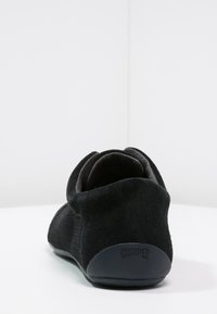 Svart slip-on sko med mjuk mockaklack, texturerade tygsidor och "Camper"-logotyp på klacken, visad bakifrån på en vit yta.
