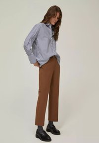 Camicia a righe blu e bianche oversize con bottoni, pantaloni marroni su misura, mocassini neri e calze grigie a coste su sfondo neutro.