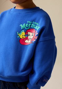 Sweatshirt bleu avec un graphisme d'Ariel et Flounder, aux couleurs vives et aux poignets côtelés. Étiquette brodée "Disney Princess" sur la manche.
