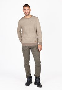 Beige gebreide sweater met een textuur, voorzien van geribde boorden en onderrand, gecombineerd met olijfgroene cargobroek met meerdere zakken en zwarte veterschoenen.