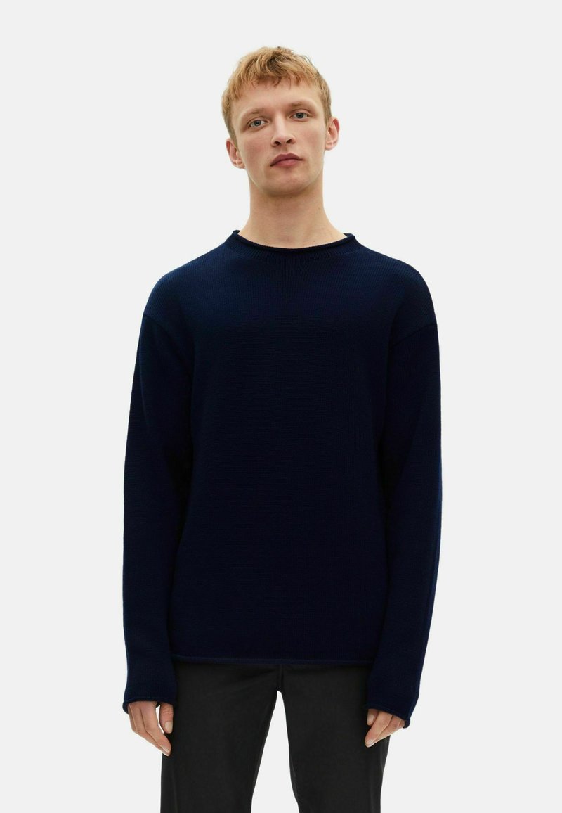 Dunkelblauer gestrickter Pullover mit rundem Ausschnitt, überschnittenen Schultern und langen Ärmeln. Der Stoff hat eine strukturierte, gerippte Optik. Schwarze Hose.