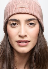 Il berretto a costine beige presenta una superficie testurizzata e una patch con la scritta "COLORFUL SIDES OF LIFE", indossato sopra capelli castani medi.